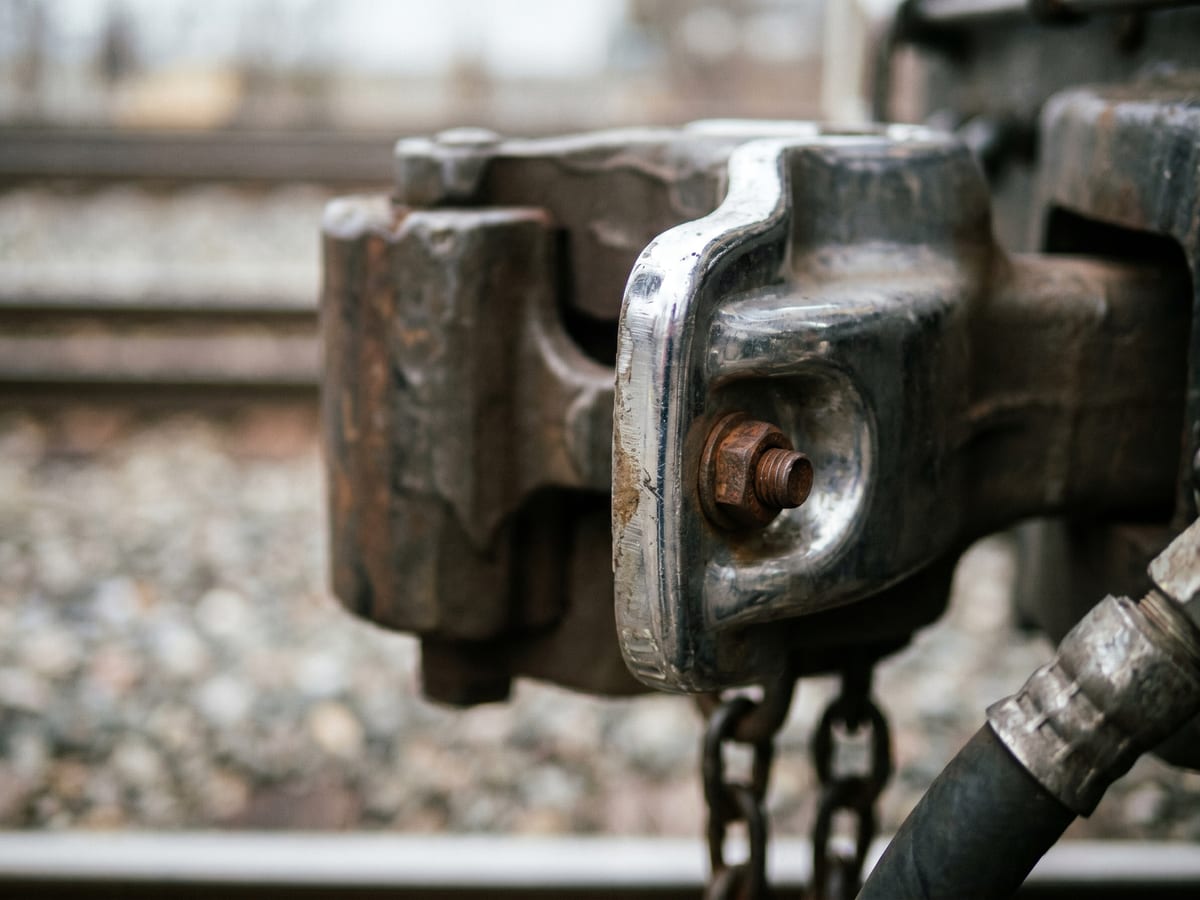 Extreme close-up of a worn metal train coupler with shallow depth of field — focus locked on the rusted bolt while the ballast and tracks behind dissolve into soft bokeh