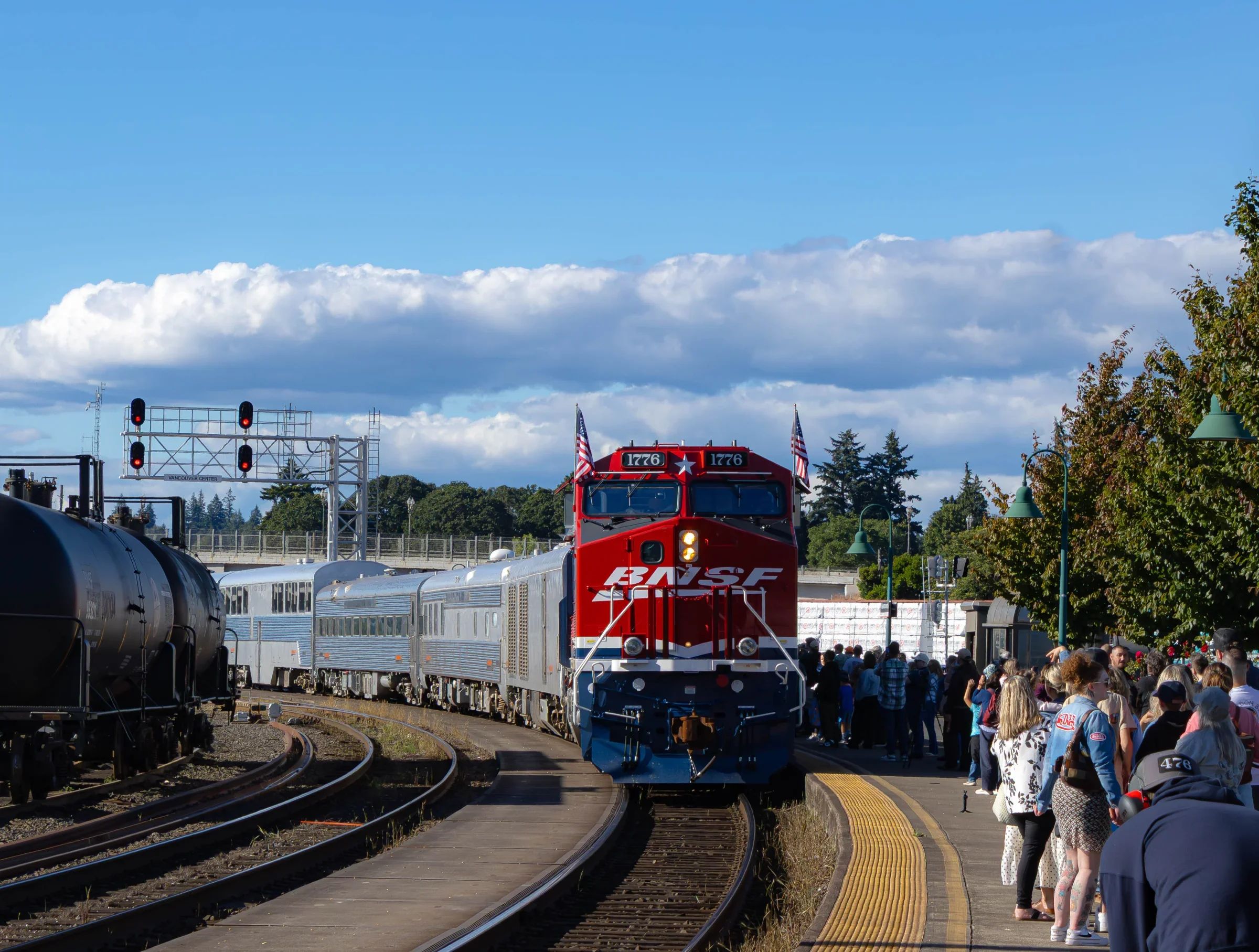 Red white and blue BNSF locomotive pulling a string of silver cars arrives at a passenger platform, greeted by a large crowd